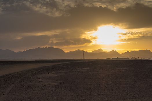 Sandy land in the foreground, rocky mountains in the background. Little clouds on the sky during the sunset.