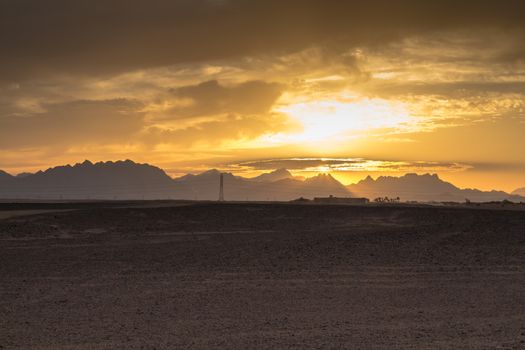 Sandy land in the foreground, rocky mountains in the background. Little clouds on the sky during the sunset.