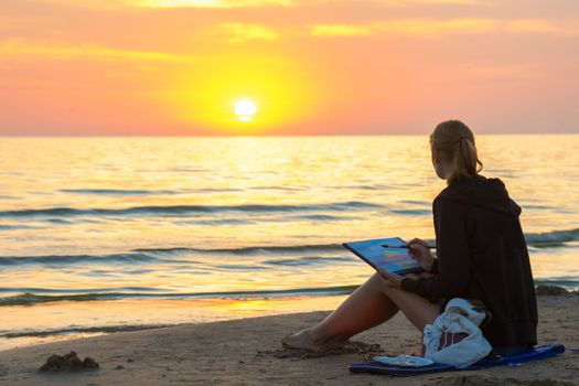 Young girl sitting on the beach and water paints watercolor landscape with sunset on a piece of paper
