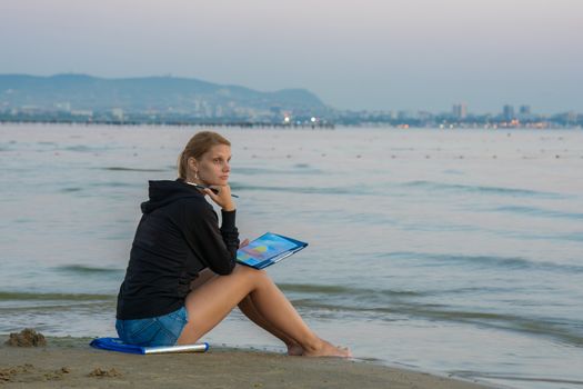 Young girl sitting on the beach and water paints watercolor landscape with sunset on a piece of paper