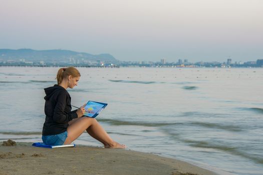 Young girl sitting on the beach and water paints watercolor landscape with sunset on a piece of paper