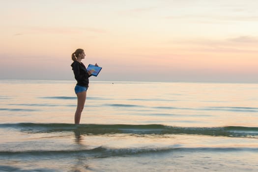 Young girl standing in the water on the beach, and draws on the colors of the landscape with sunset on a piece of paper