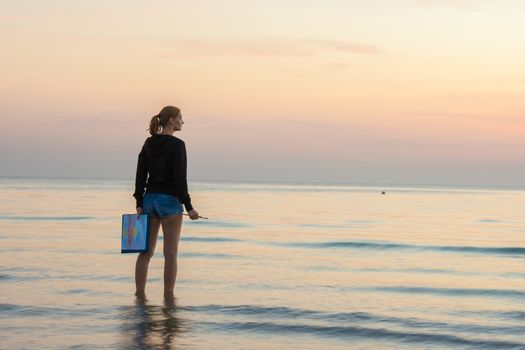 Young girl standing in the water on the beach, and draws on the colors of the landscape with sunset on a piece of paper