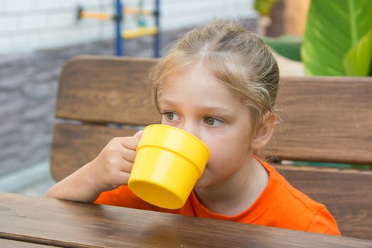 Four-year girl sitting at a wooden table on the porch and drink from the cup