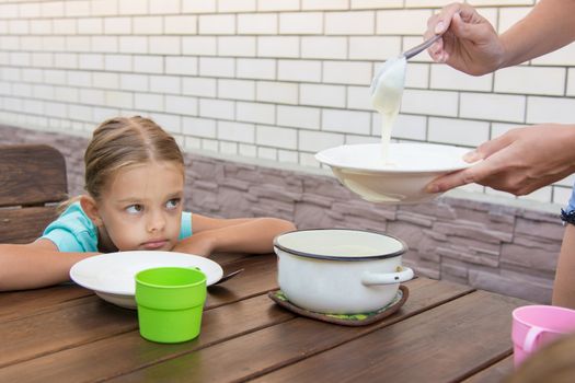 Six-year girl sitting at a table with a sour face and did not want to eat porridge for breakfast