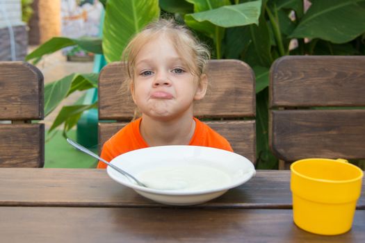 Five-year girl sits at a table with a sour face and did not want to eat porridge for breakfast