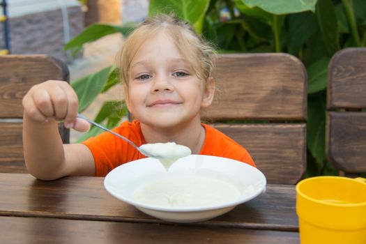 Five-year girl sits at a table on the veranda and enjoy eating porridge