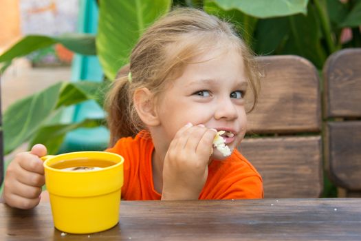 Four-year girl sitting at a wooden table on the porch, drinking a drink from the cup and eating a bun