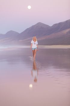 Woman walking on sandy beach in dusk leaving footprints in the sand. Beach, travel, concept. Copy space. Vertical composition.