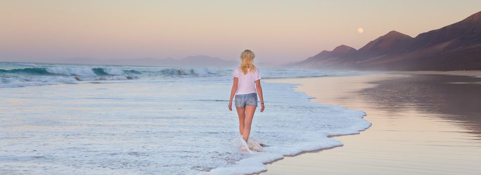 Woman walking on sandy beach in sunset. Waves sweeping away her traces in sand. Beach, travel, concept. Copy space. Panoramic composition.