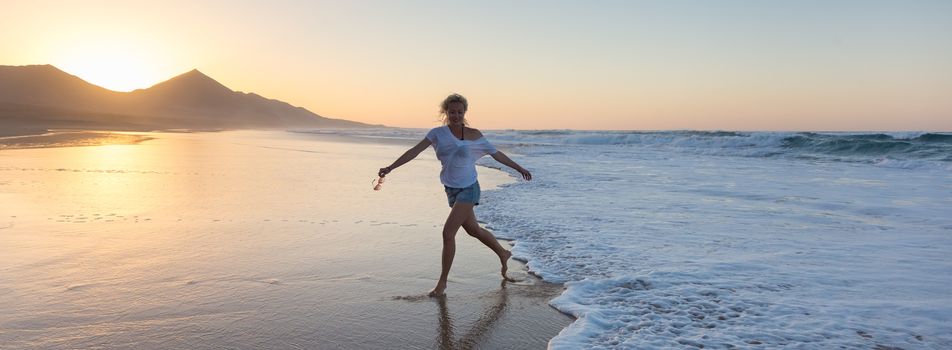 Woman having fun runing from waves on solitary sandy beach in sunset. Waves sweeping away her traces in sand. Beach, travel, concept. Panoramic composition.