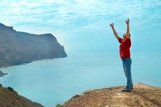 Happy man standing on the cliff with hands up looking at the sea