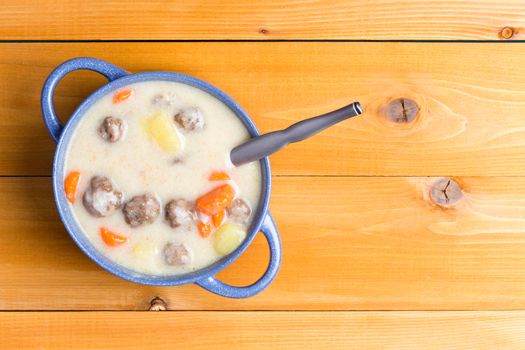 Homemade meatball soup with fresh vegetables in a creamy broth served in a blue cup or bowl on a wooden table with copy space, overhead view