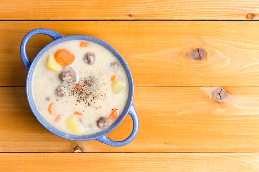 Nutritious meatball soup with vegetables garnished with ground black pepper and served in a blue bowl on a natural wood table with copy space, overhead view