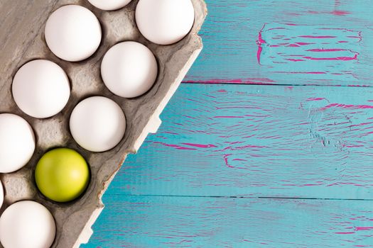 Egg box full of white Easter eggs with one single green one in a conceptual image, overhead view on a crackle painted blue table with copy space