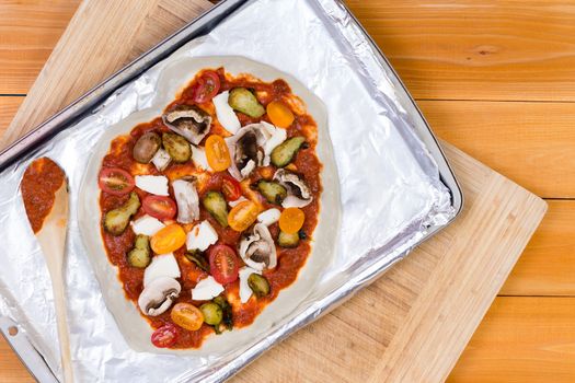 Hand tossed homemade vegetarian pizza topped with tomato paste and assorted fresh vegetables, mozzarella cheese and mushrooms ready to cook on a baking tray, overhead view