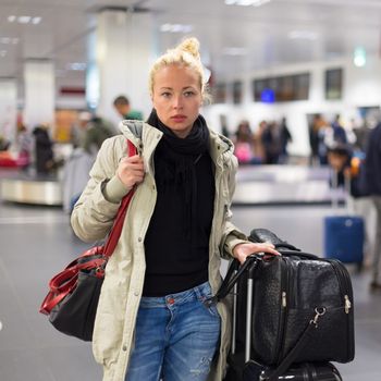 Casually dressed young female traveller walking in airport terminal hall with collected laggage after flight arrival . Blured background. Can also be used as railway, metro, bus station.