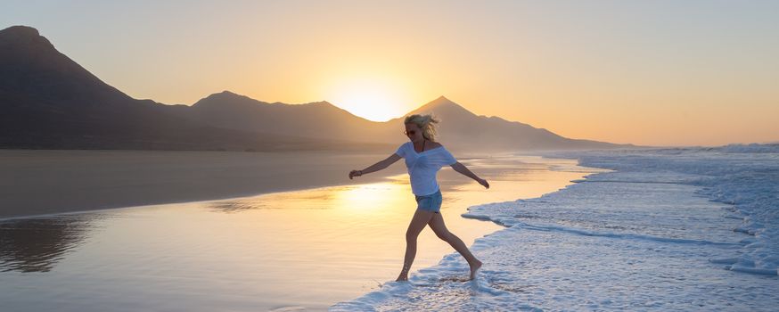 Woman having fun runing from waves on solitary sandy beach in sunset. Waves sweeping away her traces in sand. Beach, travel, concept. Copy space. Panoramic composition.