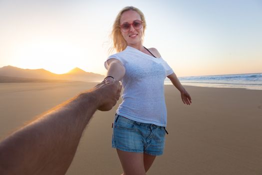 Young romantic couple, holding hands, having fun on perfect deserted beach at sunset. Shot from boyfrieds perspective. Guy looking at her beautiful carefree girlfriend.