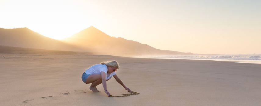 Woman drawing heart in sand on beautiful solitary Cofete beach in sunset. Love, travel concept. Fuerteventura, Canary Islands, Spain.