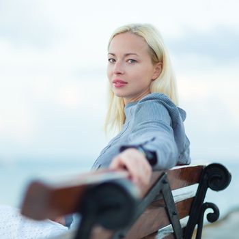 Beautiful woman sitting on a vintage wooden bench, relaxing on fresh breeze by the sea, looking at camera.