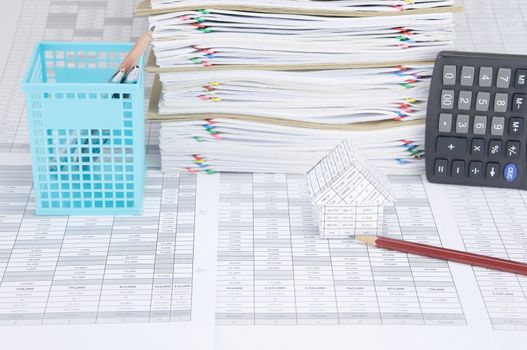 House and brown pencil on finance account have pen in blue basket and calculator place vertical beside overload of paperwork and brown envelope with colorful paperclip as background.