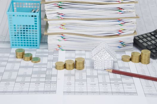 House and pencil with step pile of gold coins on finance account have pen in blue basket and calculator beside overload of paperwork and brown envelope with colorful paperclip as background.