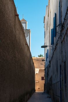 Narrow street with wall on one side and residential house with blue shutters on the other side. Tower of a mosque visible in the background. Bright blue sky.