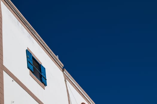 White facade of a house with opened window with blue shutter, matching with the deep blue color of the sky. Essouira, Morocco.