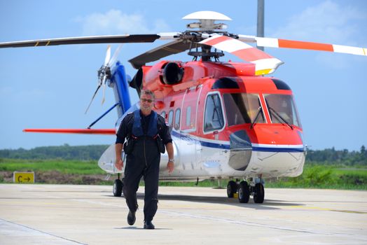 caucasian offshore helicopter pilot is walking and smiling beside of helicopter at apron