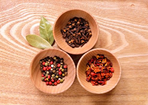 Threepepper, cloves and slices of dried peppers on a light wooden table. Next to the bowl are two of dried bay leaves.