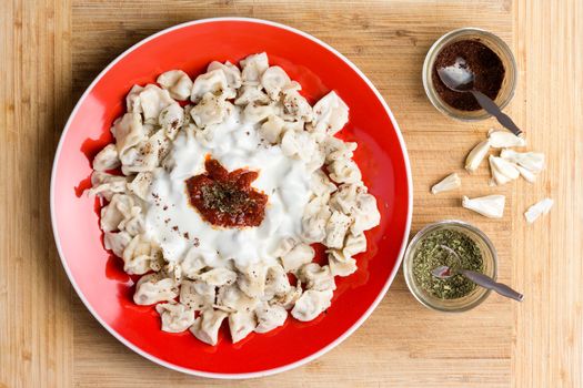 Delicious Turkish dumplings prepared and served on red plate next to seasonings in cups and chopped raw garlic