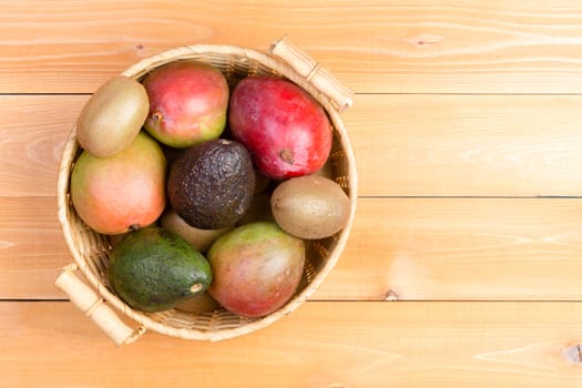 Top down view of large round basket with two handles full of avocados, mangoes and kiwi fruit over wooden background