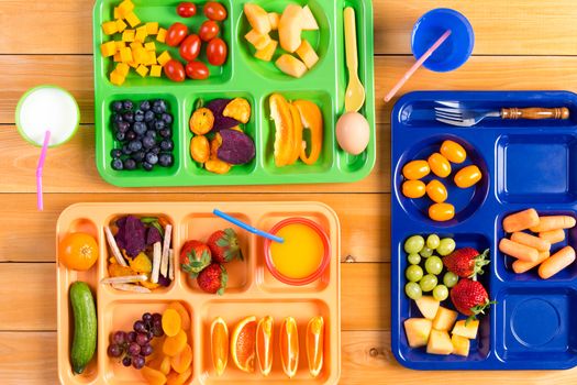 Top down view of colorful plastic lunch trays filled with slices and pieces of fruit and vegetables along side cups of milk or juice on wooden table