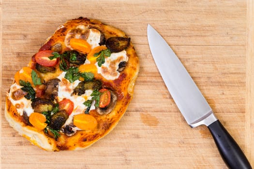 Freshly baked hand tossed vegetarian pizza with mushrooms, tomato and mozzarella cheese on a wooden cutting board alongside a kitchen knife