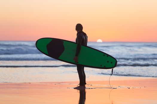 Silhouette of male surfer on the beach with the surfboard watching sunset.