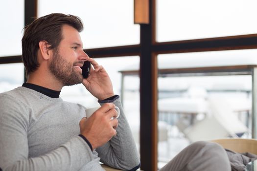 Relaxed charming man sitting in front of window with cup of morning coffee or tea near window chatting on phone.