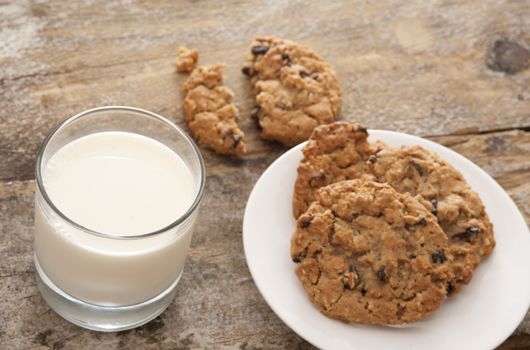 Freshly baked chocolate chip or raisin cookies in little white plate and milk in glass on wooden table