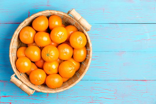 Top down first person perspective view of straw basket filled with clementine oranges over painted blue wooden table with copy space