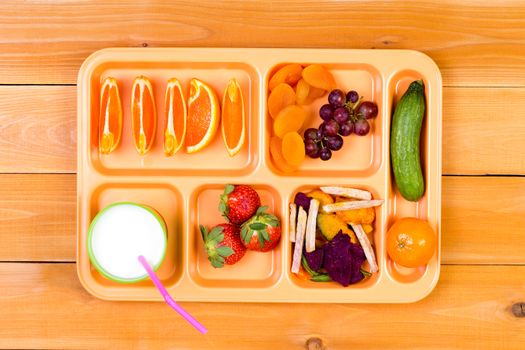 Lunchtime plastic tray filled with orange wedges, dried apricot, grapes, pickle, strawberry and other ingredients next to cup of milk over wooden table background