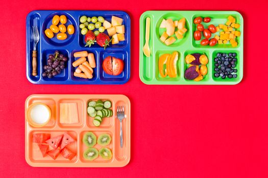 Three plastic colorful kids lunch trays filled with tomatoes, grapes, watermelon and other produce over red background with copy space