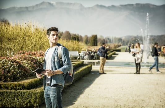 Handsome young man wearing a backpack sightseeing in a lush green autumn park, standing alongside a canal with swans holding a map or brochure, looking into the distance as though lost