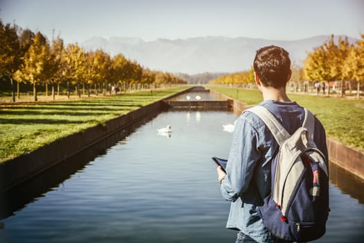 Handsome young man wearing a backpack sightseeing in a lush green autumn park, standing alongside a canal with swans holding a map or brochure, looking into the distance as though lost