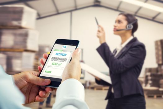hand holding smartphone against warehouse manager wearing headset checking inventory 