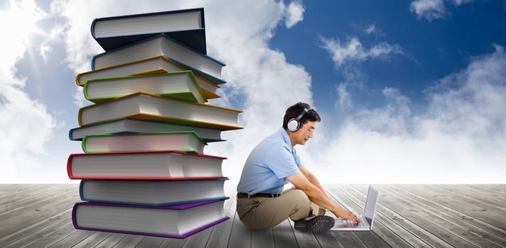 Side view of man listening music while using laptop against stack of books against sky