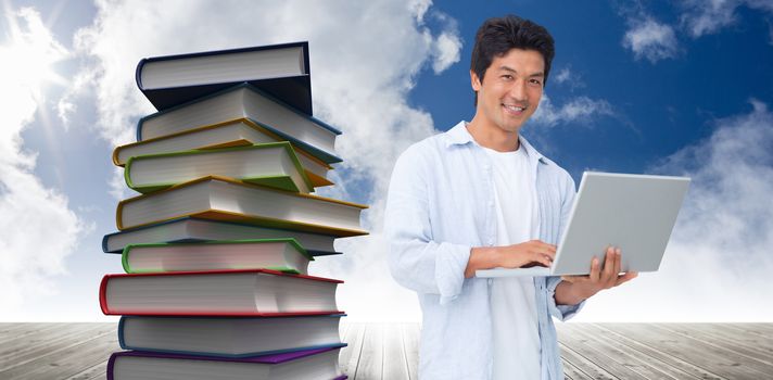 Smiling male with his laptop against stack of books against sky