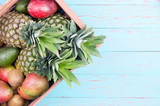 Rectangular cardboard box of pineapples, mangoes, avocados and kiwi fruits over wood planks in blue with copy space