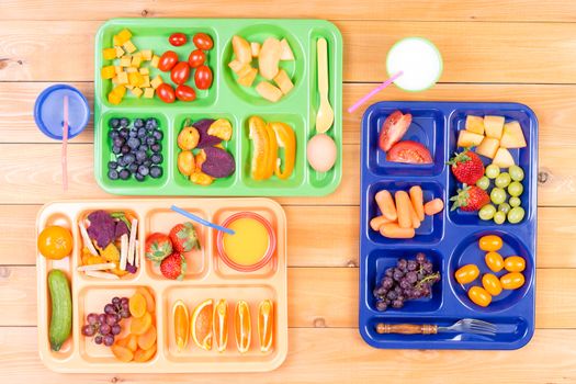 Top down view of colorful plastic lunch trays filled with delicious pieces of fruit and vegetables along side cups of milk or juice on wooden table
