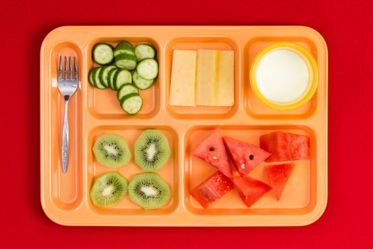 Top down perspective view on bright plastic child size lunch tray with fork, cucumber, cheese, kiwi, watermelon slices and little cup of milk