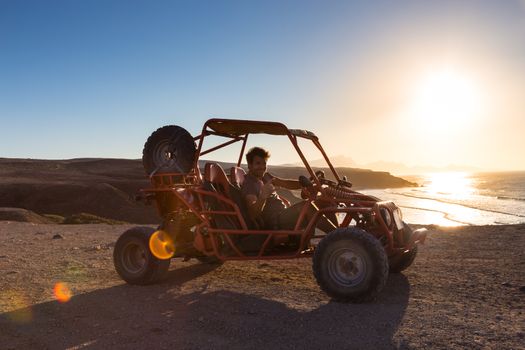 Active man driving quadbike on dirt road by the sea in sunset showing rocking sign to the camera.